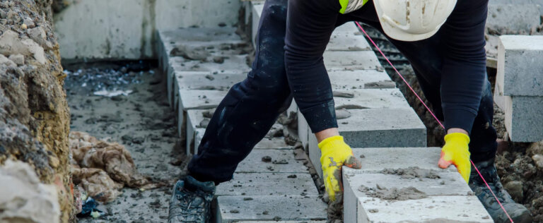 Hard working bricklayer laying concrete blocks on top of concrete foundation ground beam on new residential housing site. Fight housing crisis by building more affordable houses concept