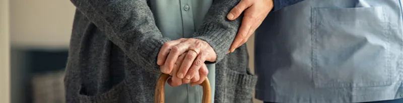 Elderly woman walking with cane, being assisted by a nurisng home worker.